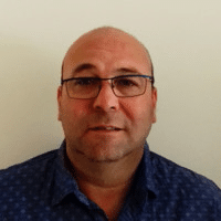 Homme avec lunettes pose devant un mur blanc.